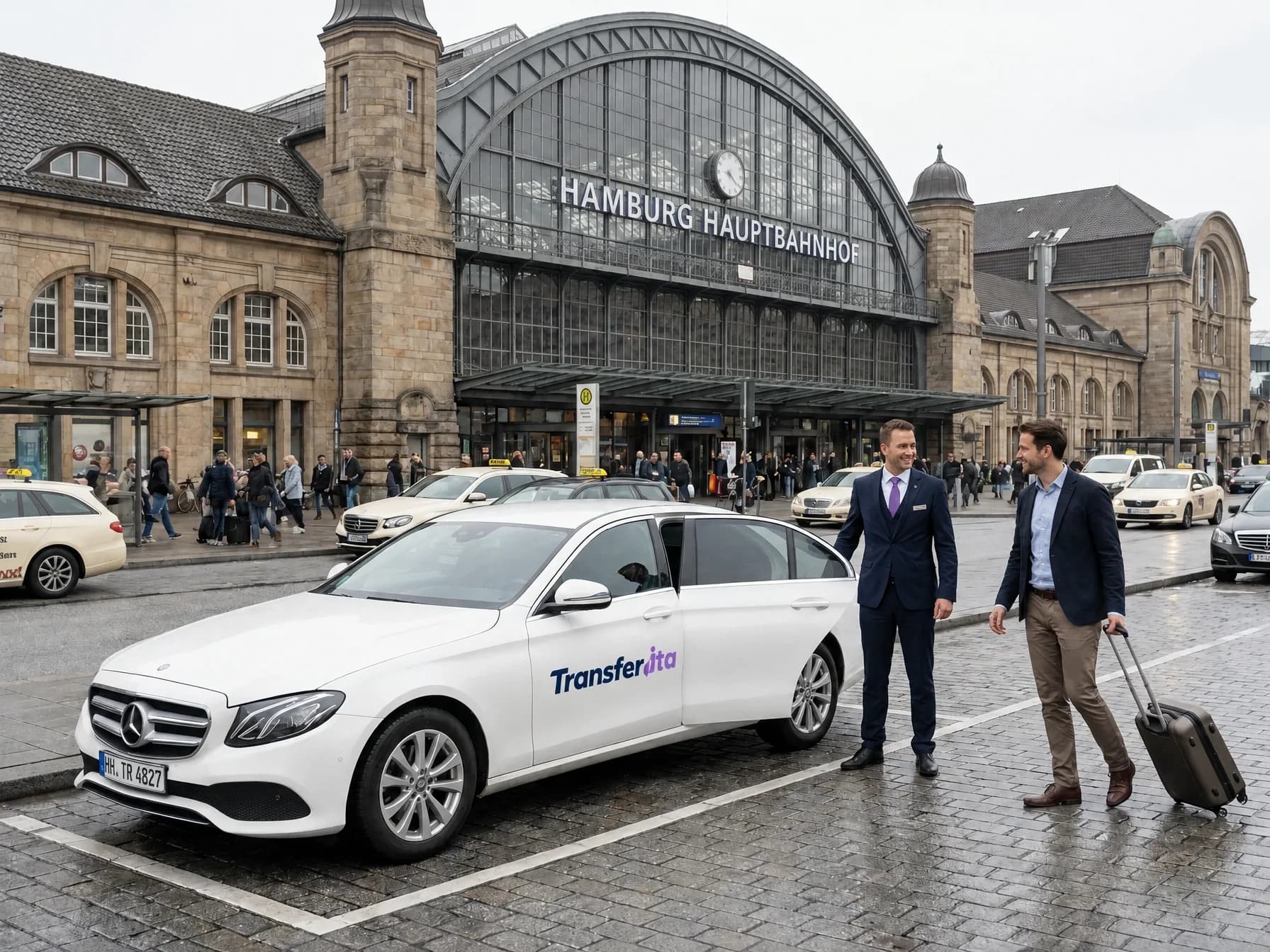 Fahrer im Anzug an einem weißen Wagen mit Transferita-Schriftzug vor dem Hamburger Hauptbahnhof, ein Fahrgast kommt mit Rollkoffer dazu