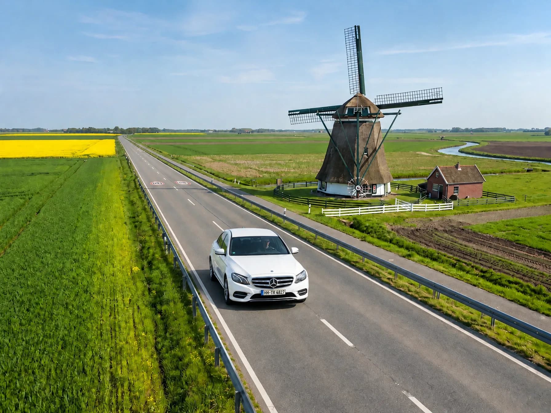 Weiße Limousine auf einer Landstraße zwischen grünen Wiesen und einem gelben Rapsfeld, daneben eine Windmühle mit Reetdach und ein kleines Backsteinhaus
