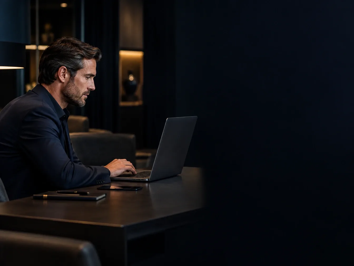Mann im dunklen Sakko arbeitet abends an einem Tisch am Laptop
